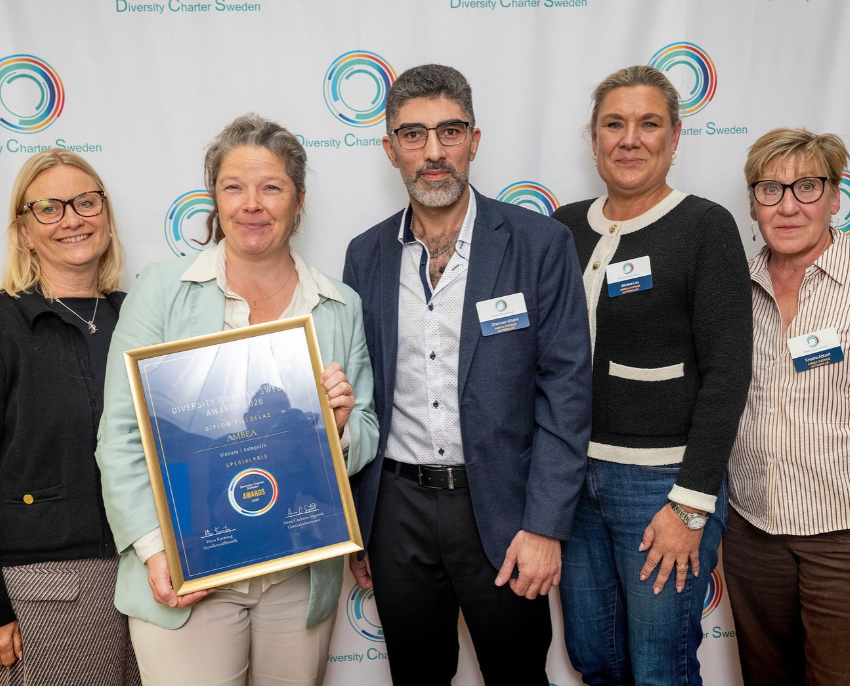 Fem personer står framför en vägg med Diversity Charter Swedens logotyp. En person håller upp ett inramat diplom där Ambea utses till vinnare av Specialpriset i Diversity Charter Sweden Awards 2026.